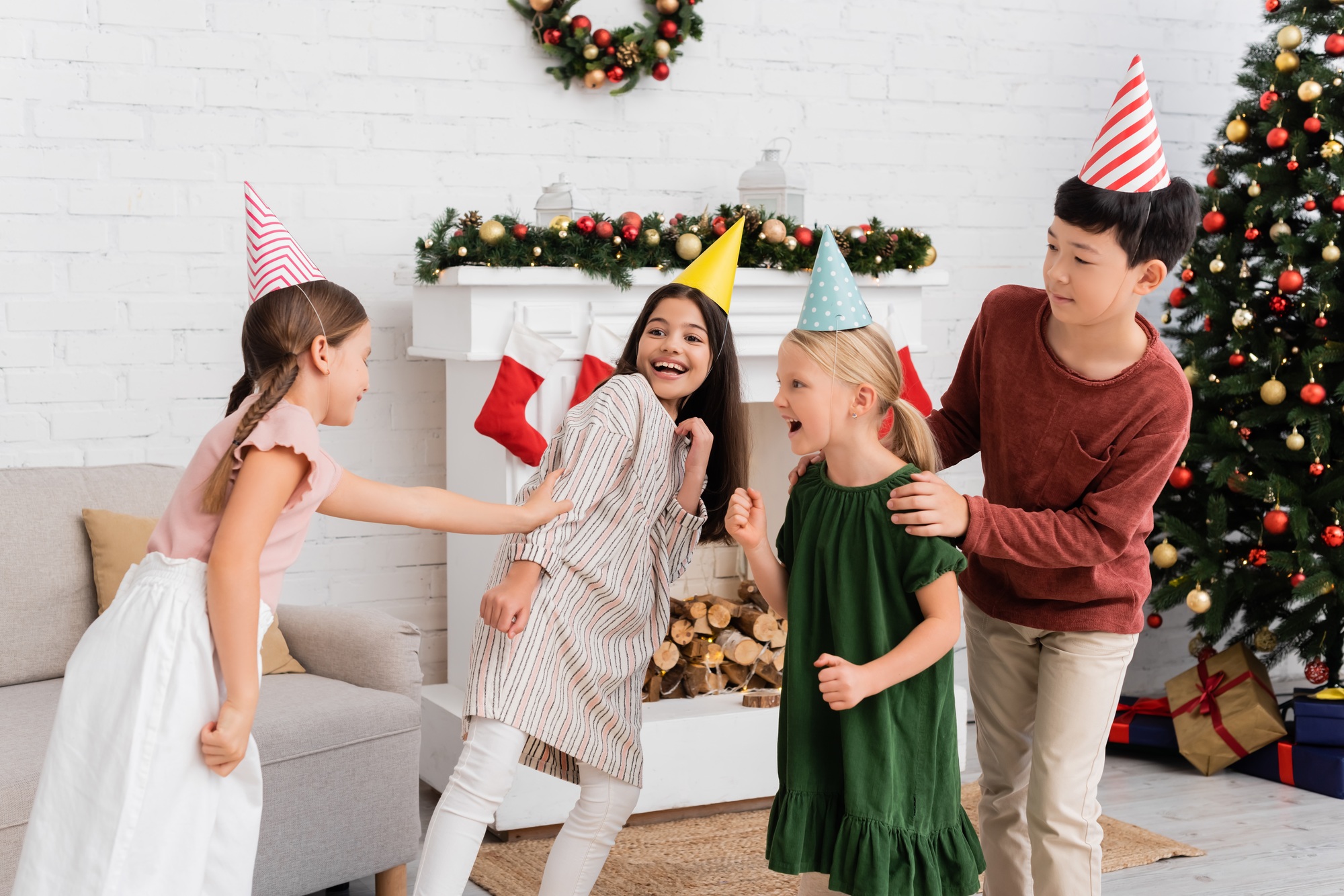 Cheerful interracial kids in party caps playing tag during birthday party near christmas decor at